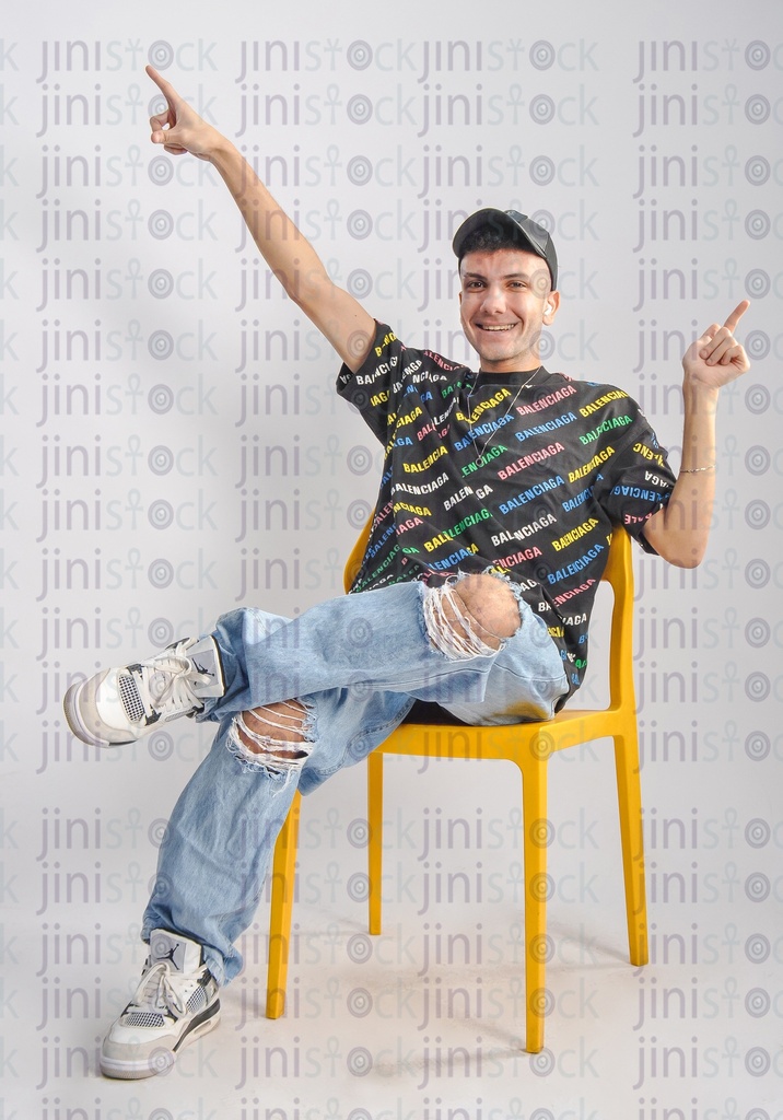 a cool young man wearing a cap, t-shirt, and jeans, and sitting on a chair and listening to music and pointing