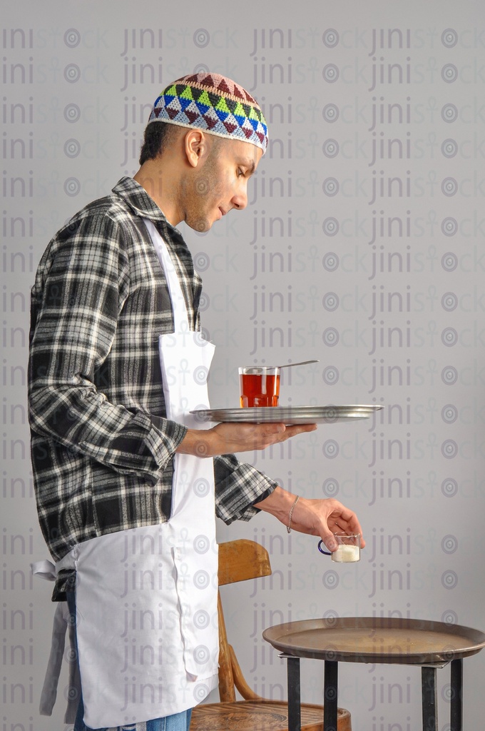 a man holding a tray of tea and holding the sugar in his hand wearing work clothes for a coffee shop or café.
