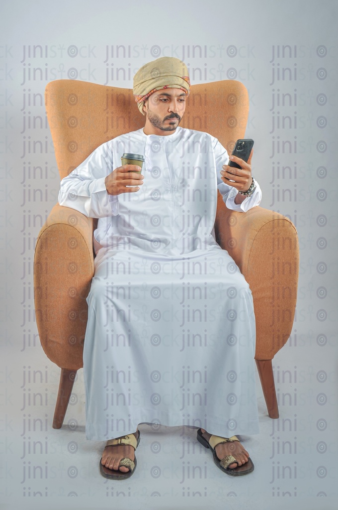 An Omani man from the Gulf wearing Omani uniform sitting on a chair, holding a mobile phone and texting or browsing the offers via mobile applications, and he is holding a cup of coffee or tea.
