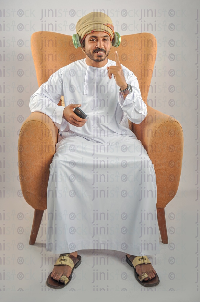رجل عماني من الخليج يرتدي الزي العماني ويجلس على كرسي ويستمع إلى الأغاني.