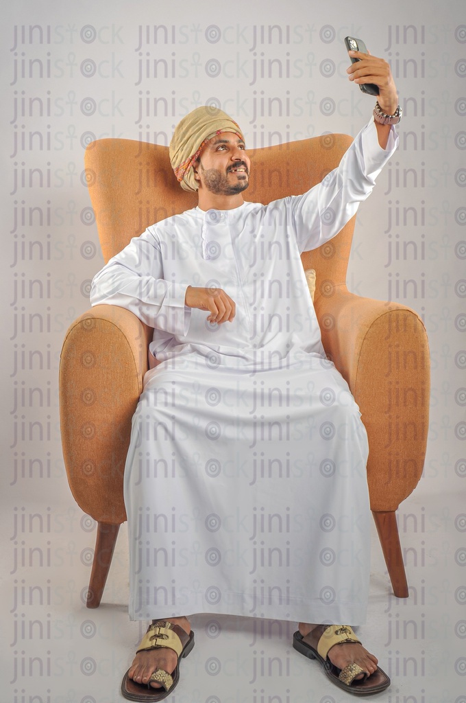 An Omani man from the Gulf wearing Omani uniform sitting on a chair and taking a selfie photo or making a video call