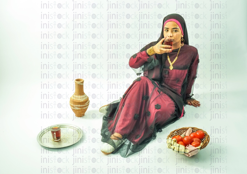 امرأة مصرية نوبية ترتدي ثوبا نوبيا وحجاب على رأسها تجلس على الأرض وتشرب كوبا من الشاي. يوجد طبق من الطماطم ، قله " فخار" ، وكوب من الشاي على صينية على الأرض