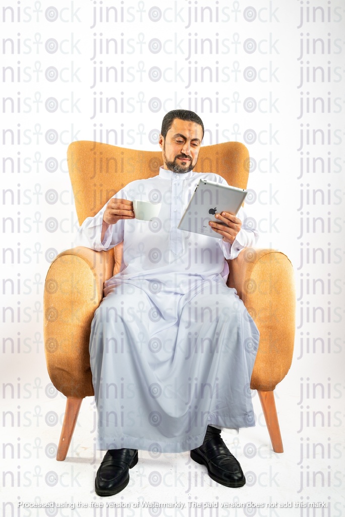 رجل سعودي خليجي من الخليج يجلس على كرسي ينظر إلى جهاز الآيباد الخاص به ويشرب القهوة في الكوب