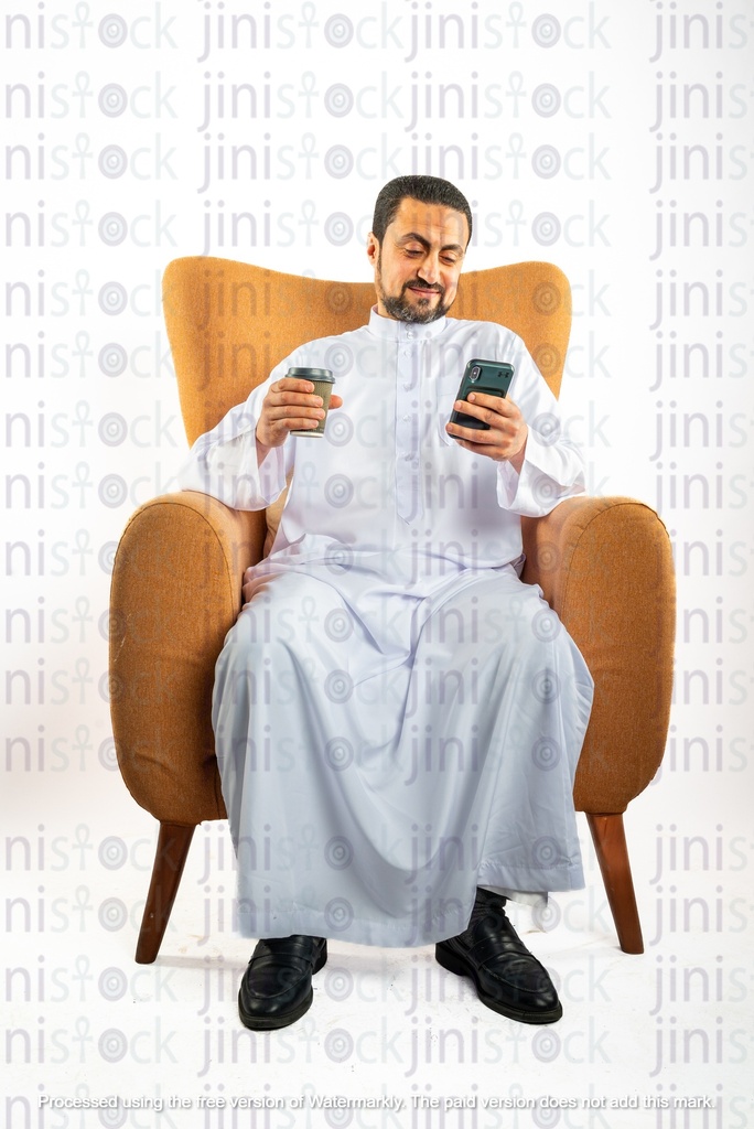 رجل سعودي خليجي يجلس على كرسي ويتحقق من هاتفه المحمول ويحمل كوب قهوة من ناحية أخرى