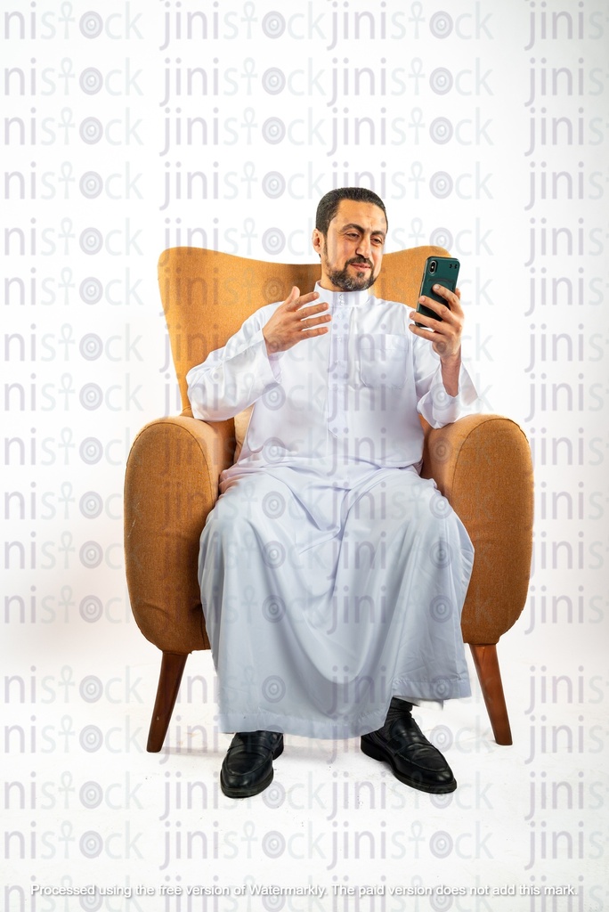 رجل سعودي خليجي يجلس على الكرسي سعيدًا وينظر إلى هاتفه المحمول