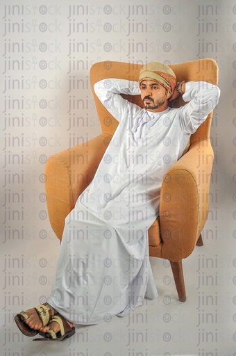 An Omani man from the Gulf wearing Omani clothes sitting on a chair putting his hands behind his head and thinking of something