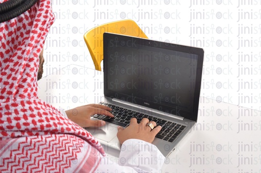 لقطة من فوق الكتف لشخص سعودي أو خليجي أو إماراتي أو رجل من الخليج يعمل على جهاز كمبيوتر محمول في مكتبه.