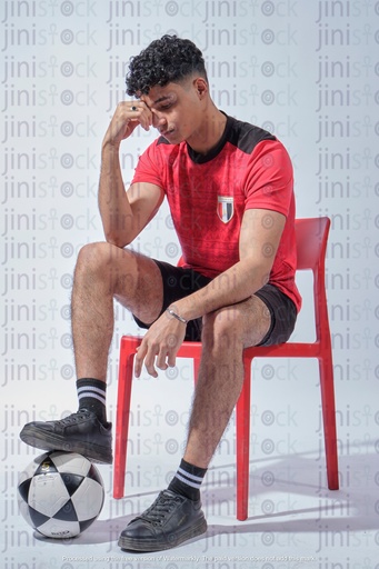 young Egyptian man sitting on a chair wearing the Egyptian national team shirt sad