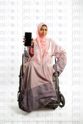 امرأة خليجية سعودية من الخليج على كرسي متحرك وتعاني من إعاقة وتحمل هاتفاً محمولاً وتشير به إلى الكاميرا.