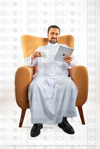 رجل سعودي خليجي يجلس على الكرسي ويتحقق من جهاز الآيباد الخاص به ويشرب الشاي