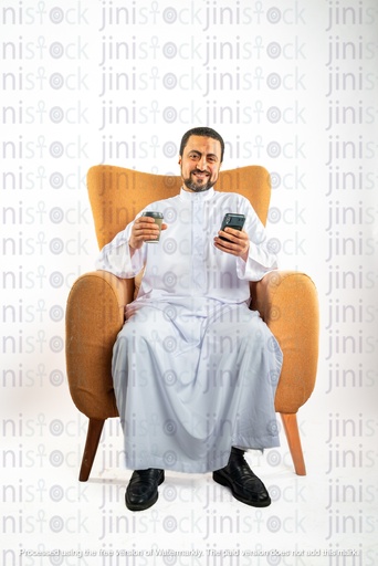 رجل سعودي خليجي سعيد من الخليج يجلس على كرسي يحمل كوب قهوة وهاتفًا محمولًا