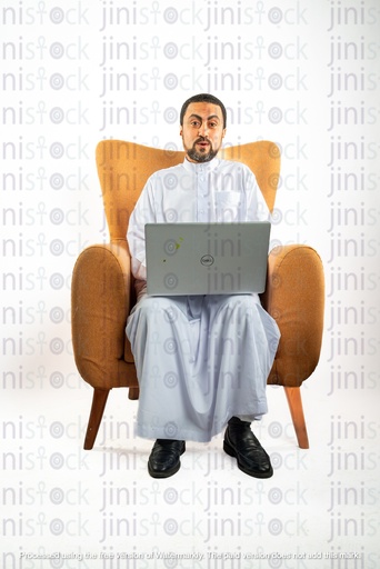 رجل سعودي خليجي من الخليج يجلس على كرسي يعمل على الكمبيوتر المحمول الخاص به وهو متفاجئ